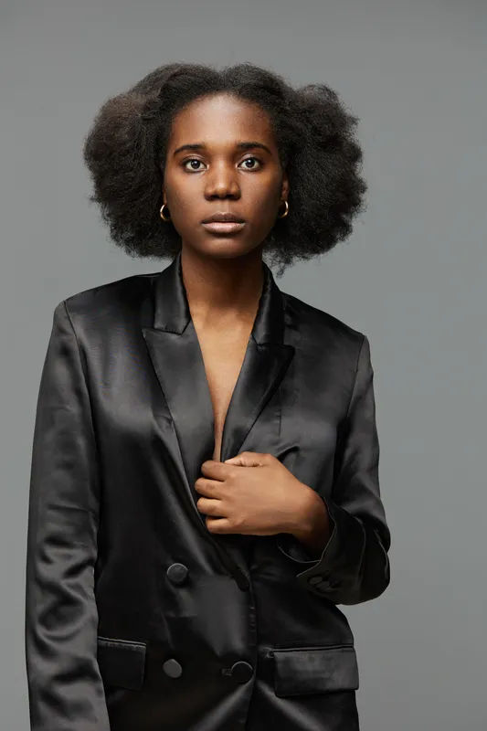 Portrait of a confident Black woman wearing a black satin blazer, showcasing natural hair against a minimalist gray background, embodying modern elegance and strength