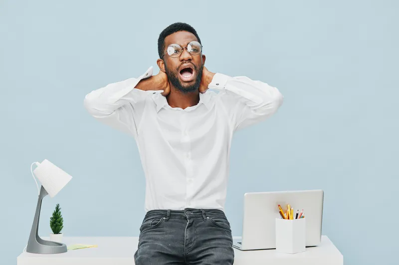 Frustrated young man with glasses, white shirt, and dark jeans stands behind a desk on a light blue background, clutching his head in stress. Office space work stress concept.