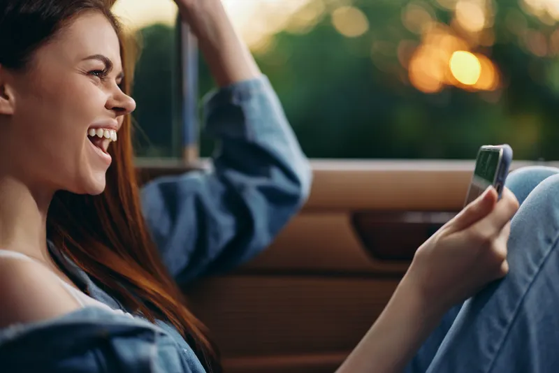 Joyful young woman laughing while using a smartphone in a car, dressed casually in denim, enjoying a sunny day outdoors with a vibrant green background.