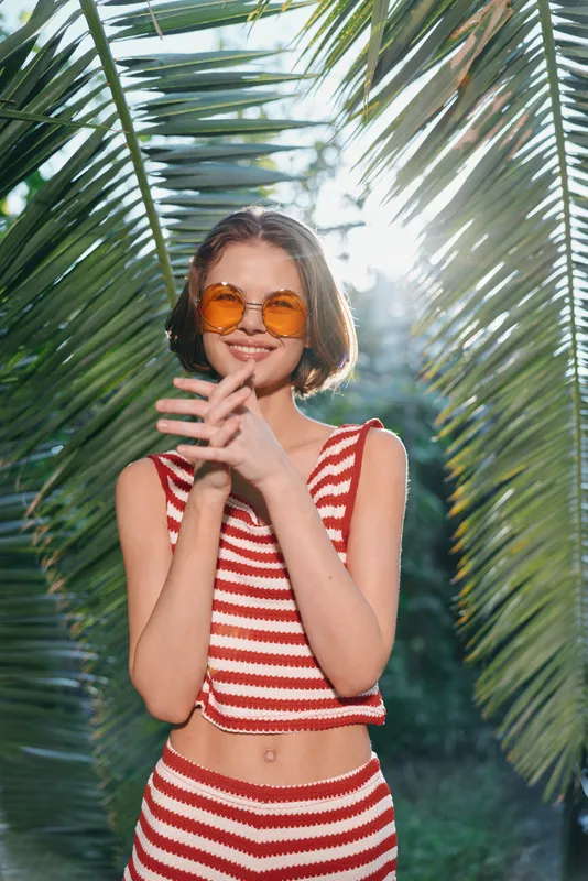 A cheerful young woman stands outdoors among tropical plants, wearing a red striped crop top and orange sunglasses. She smiles warmly, radiating sunshine, confidence, and carefree summer energy in a