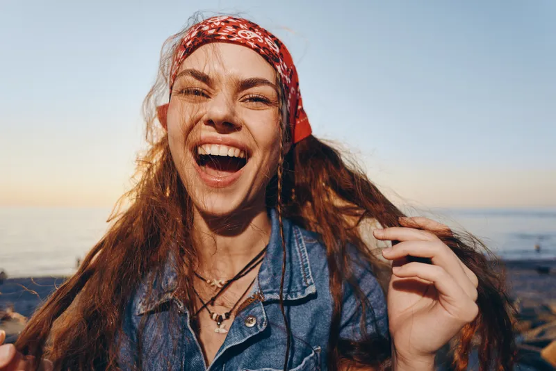 Lifestyle woman in modern boho-western denim street style laughing joyfully with film color tones, carefree and happy at sunset by the sea.