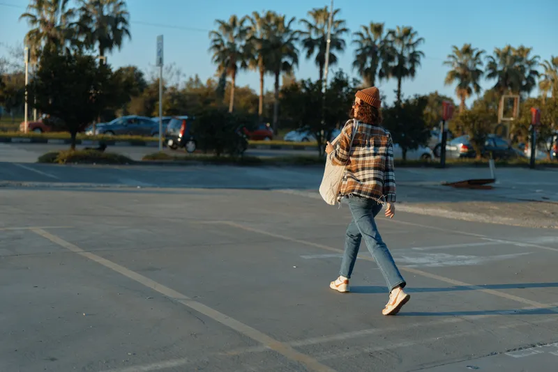 woman walking in a parking lot with urban palm backdrop, wearing plaid coat and jeans and carrying a bag at golden hour glow. Authentic candid lifestyle moment with mindful living mood.