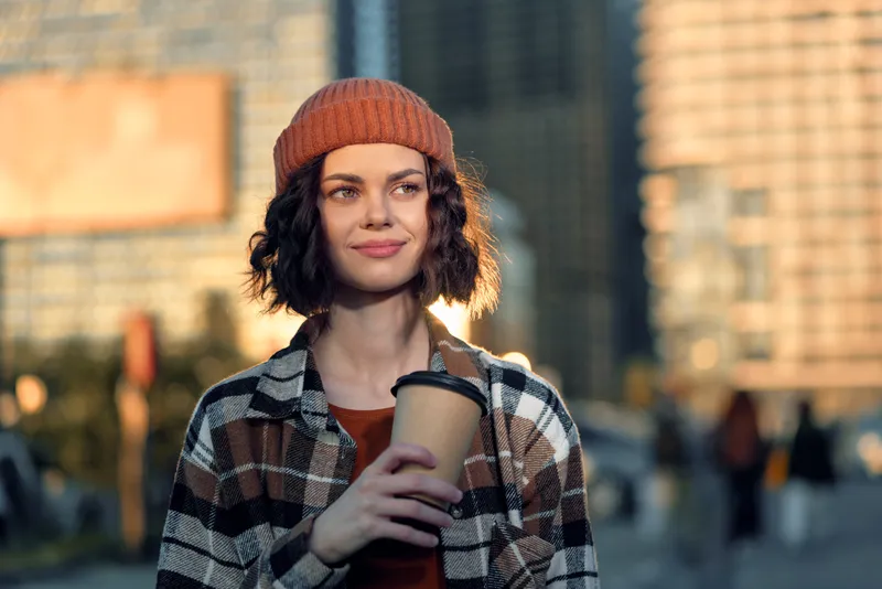 Woman holding coffee in beanie, urban portrait at morning with candid smile; authenticity and candid lifestyle captured in golden hour glow for mindful living and emotional storytelling.