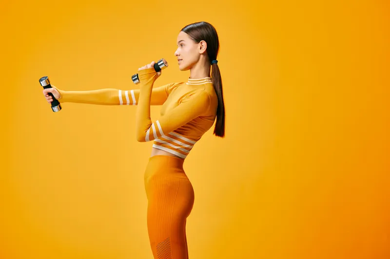 Fit woman in orange activewear lifts small dumbbells in a bright studio, side profile pose showing strength and precise form against a clean monochrome background.
