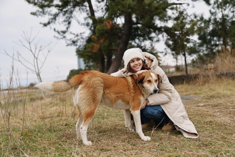 Dog and woman sharing a gentle hug in an open autumn field, bundled in a warm coat, scarf and boots. Soft natural light highlights their bond, calm mood, and peaceful outdoor moment.