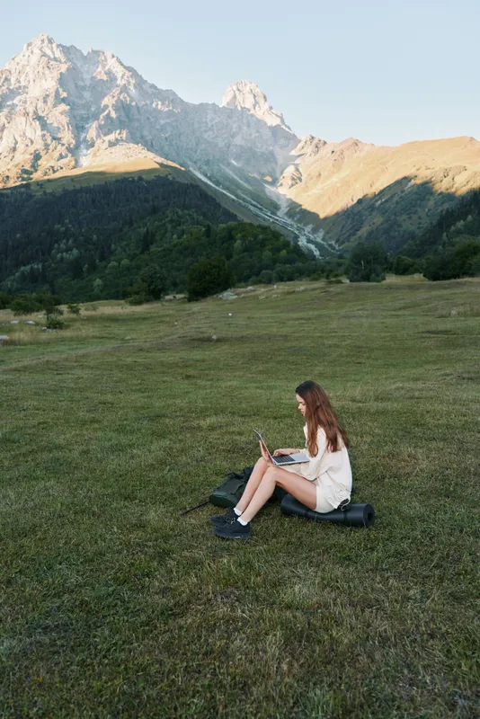A relaxed young woman sits on a grassy meadow with a laptop, surrounded by rugged mountains and blue sky, working outdoors in a peaceful, scenic setting.