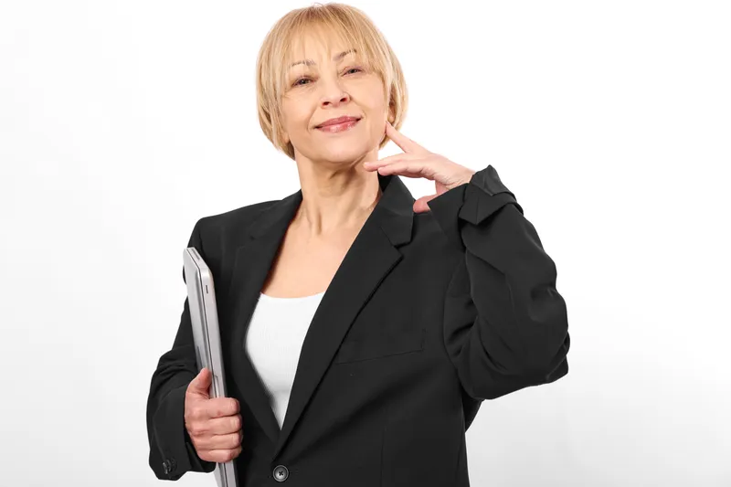 Senior woman in business blazer, holding smartphone for communication, studio portrait, smile, raised hand gesture, elegant mature look, white background