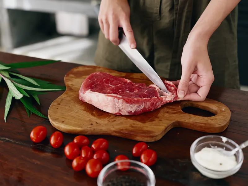 Женщина с ножом в руке нарезает свежее стейковое мясо для жарки на кухне