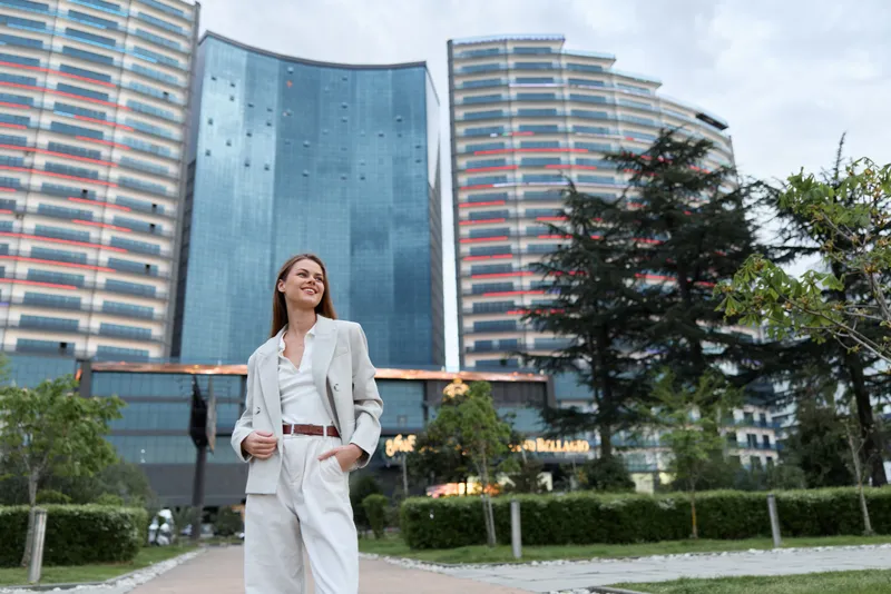 confident woman in white suit walking in modern urban business district with skyscrapers and greenery under cloudy sky