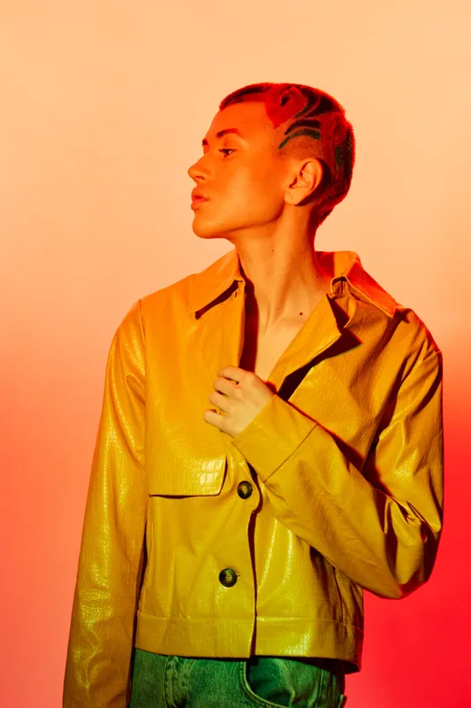 Young woman with short patterned hair wearing a yellow jacket, posing confidently against an orange background, showcasing trendy fashion and bold style.