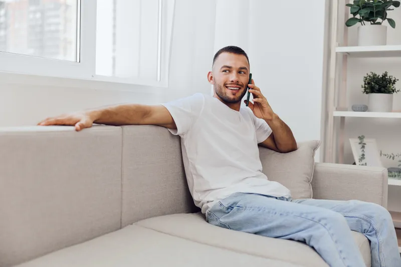 Smiling man talking on the phone with a relaxed posture, showcasing modern home decor, bright ambiance, and positive energy, ideal for lifestyle concepts.