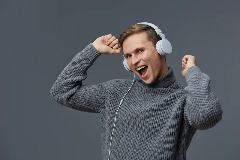 Young Caucasian man wearing gray sweater and white headphones smiling and dancing alone isolated on plain gray background studio portrait mental health and gadgets concept