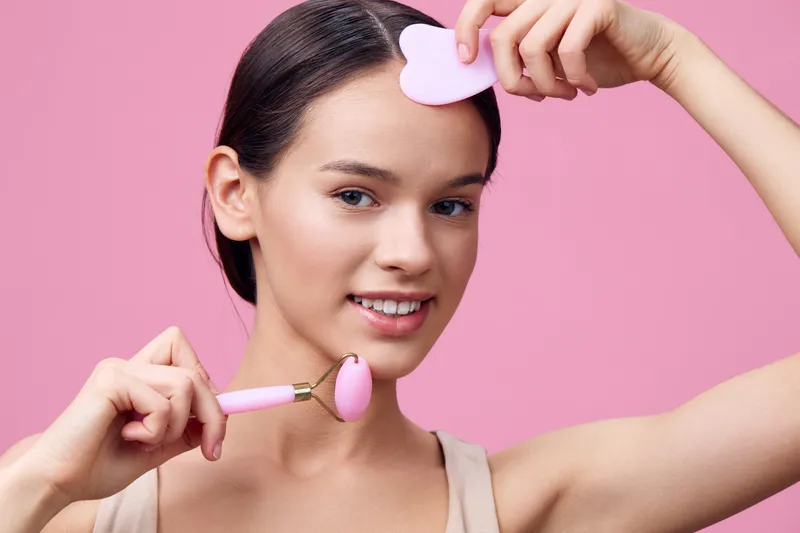 Young female model using a pink facial roller for skincare on a pink background, promoting beauty and wellness with a fresh, joyful expression
