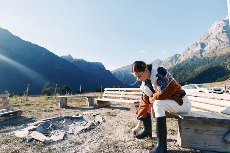 Woman in warm coat and boots sits on a wooden bench in nature by alpine mountains, drawing in the dirt near a campfire pit, enjoying solitude, scenic valley view and crisp sunlight.