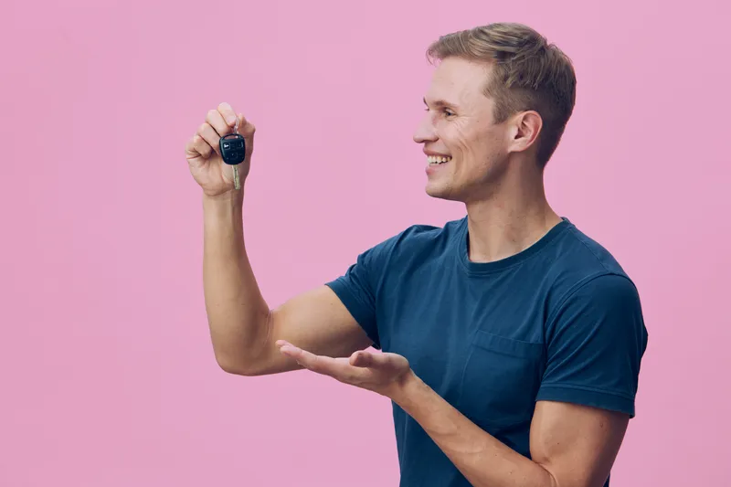 Smiling man in casual blue t-shirt holding car key, pointing to it with enthusiasm, isolated on pink background, studio portrait, lifestyle concept, happiness, success, automobile ownership, casual st