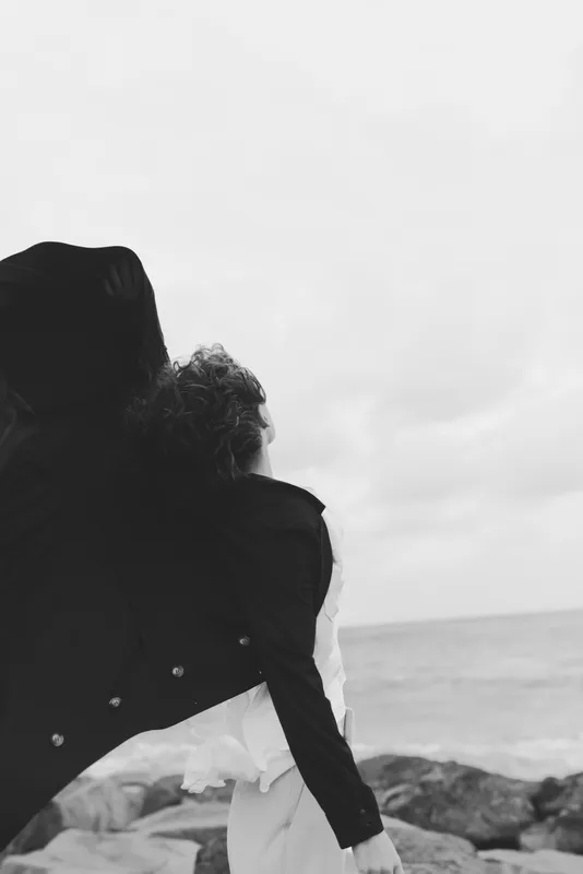 Couple embracing on a windy beach, back view in monochrome, ocean horizon in distance, capturing connection, tenderness and quiet romance against the coast, timeless coastal moment