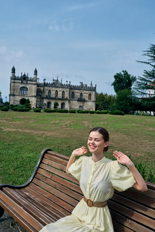 Woman smiling on a wooden bench in a sunny park with a grand castle in the background, showcasing outdoor fashion, leisure, and scenic countryside atmosphere.