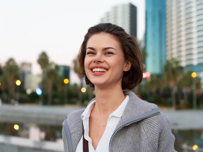 A cheerful woman stands outdoors in an urban setting, smiling brightly as she looks toward the camera, conveying confidence and approachability, energy and optimism in a casual professional scene.
