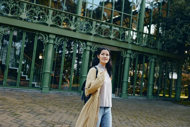 A woman wearing a beige coat and backpack stands near a green ornate railing in a park, posing for a casual outdoor shot with confident expression.