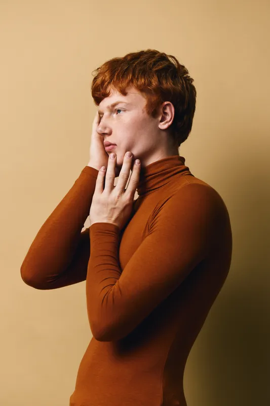 Young man with red hair and fair skin wearing brown turtleneck poses thoughtfully against beige background. Portrait side view with hand on face shows emotion and style.
