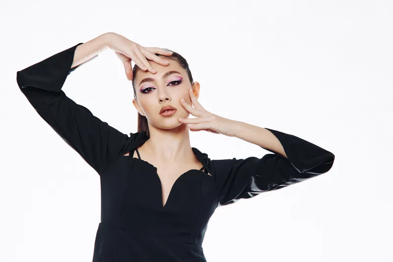Fashion model in a black dress poses with expressive hand gestures against a plain white background. The bold eye makeup emphasizes creative beauty styling and elegance.
