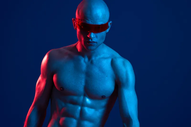 Muscular man with blue body paint and red sunglasses striking a strong pose, showcasing a captivating blend of colors and light in a vivid setting.