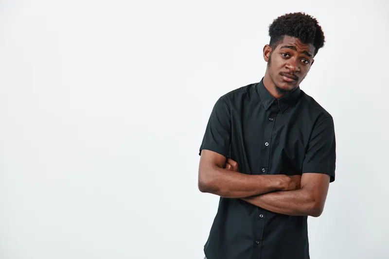 Young African American man with dark skin and curly hair wearing black shirt standing with crossed arms on white background looking at camera with thoughtful expression.