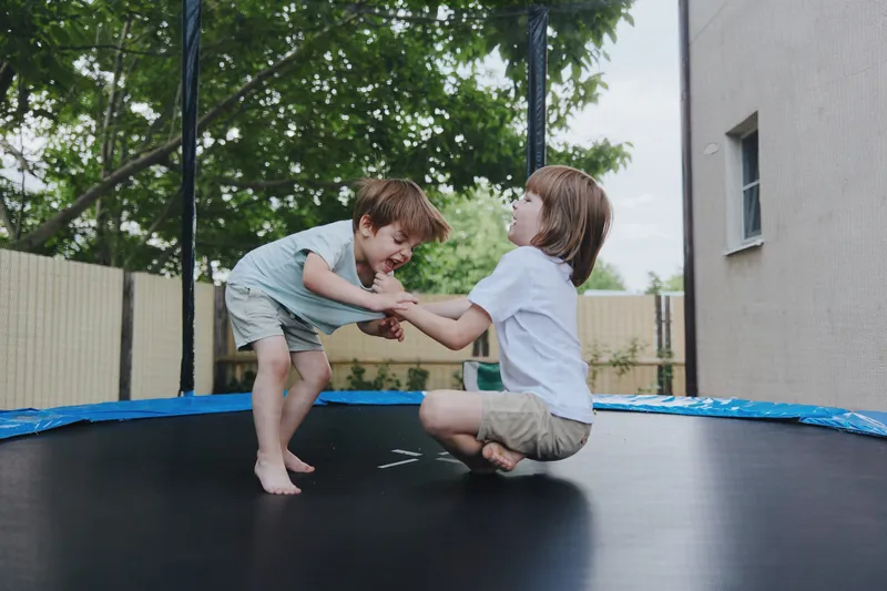 Two joyful children jumping together on a trampoline, showcasing excitement and playfulness in a sunny backyard environment.