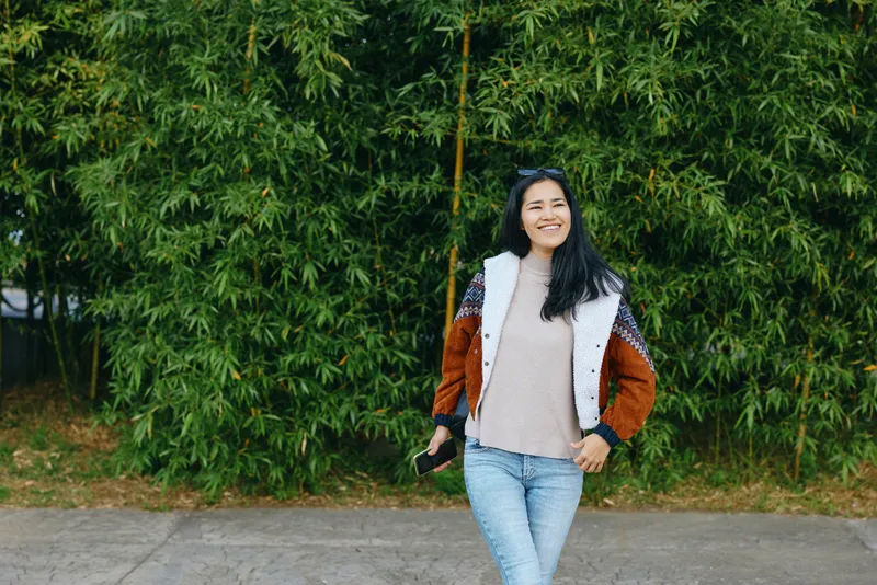 Woman outdoors with a camera, wearing a cozy jacket and jeans, walking along a leafy backdrop of bamboo, showcasing casual fashion, natural light, and a cheerful mood