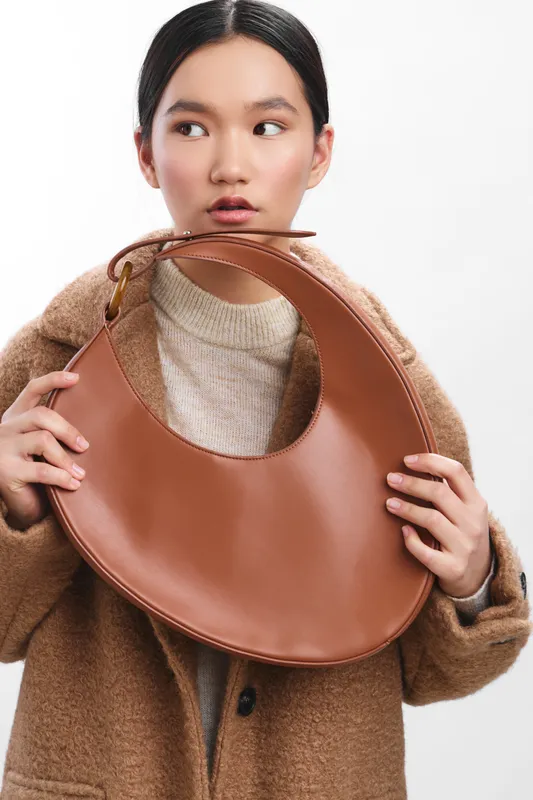 Asian woman wearing a warm coat holds a circular brown handbag near her face, looking to the side with a thoughtful and stylish expression.