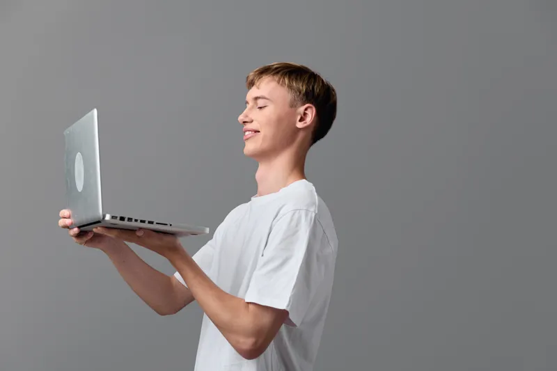 Young man holding a laptop, smiling, presenting a tech gadget for learning, remote work and digital communication in a casual studio setting, bright clean background