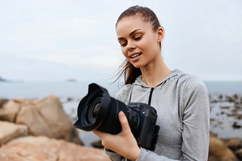 Young female photographer in sporty attire examines camera by the ocean, showcasing passion for photography in a serene coastal setting