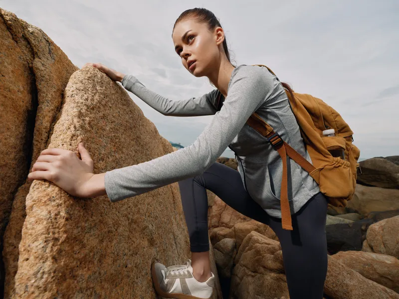 Young female hiker climbing on rocky terrain, wearing sport attire and a backpack, showcasing determination and adventure in natural outdoor setting