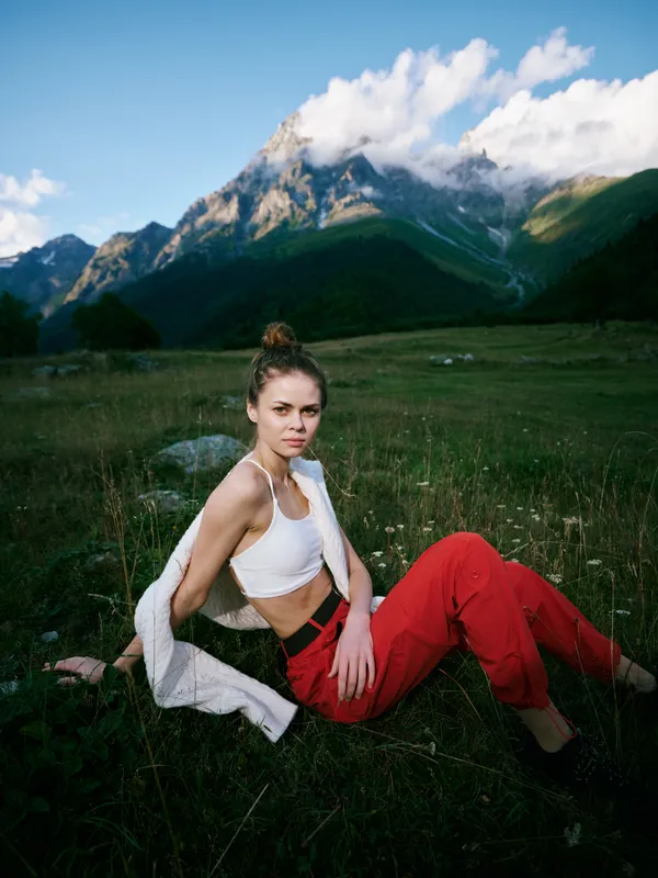 Woman sitting on a grassy meadow with mountains in the background, wearing a white top and red pants, enjoying outdoor nature, calm scenery, and fresh air.