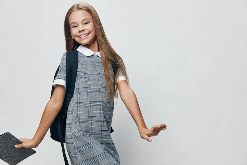 Girl, child, school, backpack and smile posing for camera on white background. Happy kid with long hair in dress holding book for education and learning at school or academy.