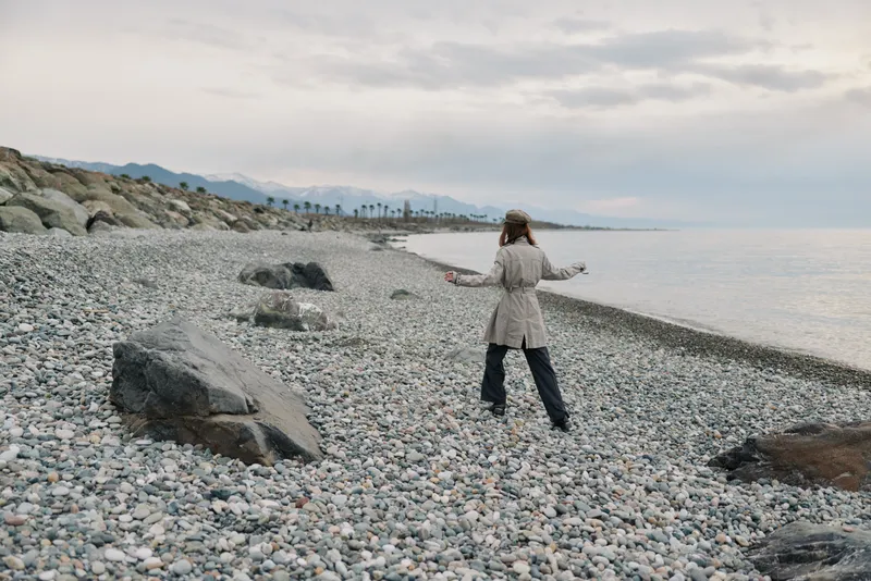 A woman in a beige coat walks alone on a rocky beach near the calm sea under a cloudy sky. The scene conveys solitude, nature, and peaceful outdoor exploration.