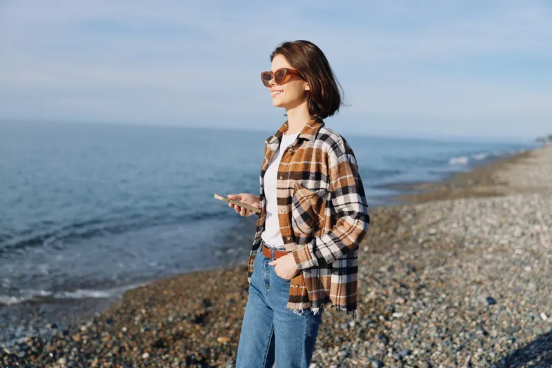 Woman on a pebble beach wearing plaid shirt, jeans and sunglasses, strolling along the coastline with a smartphone in hand under a clear blue sky.