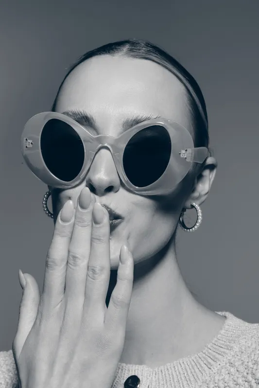 Stylish white woman with fair skin wears large round sunglasses and hoop earrings, covering mouth with hand. Concept of fashion, elegance, and mystery.