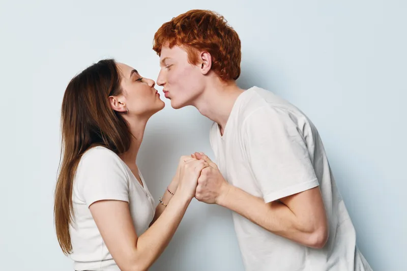 Couple in white t shirts holding hands and leaning for kiss on grey background. Young man and woman close together with eyes closed for love and affection moment at studio.