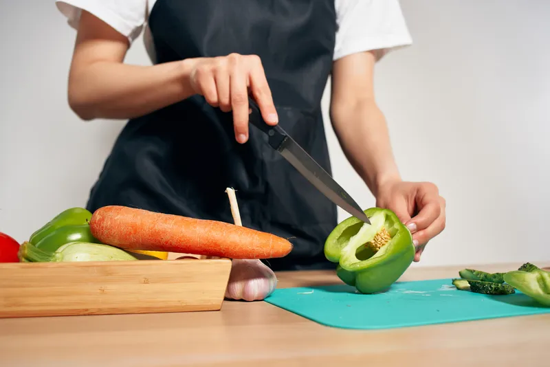 Женщина в черном фартуке нарезает овощи на кухне, домохозяйка готовит еду