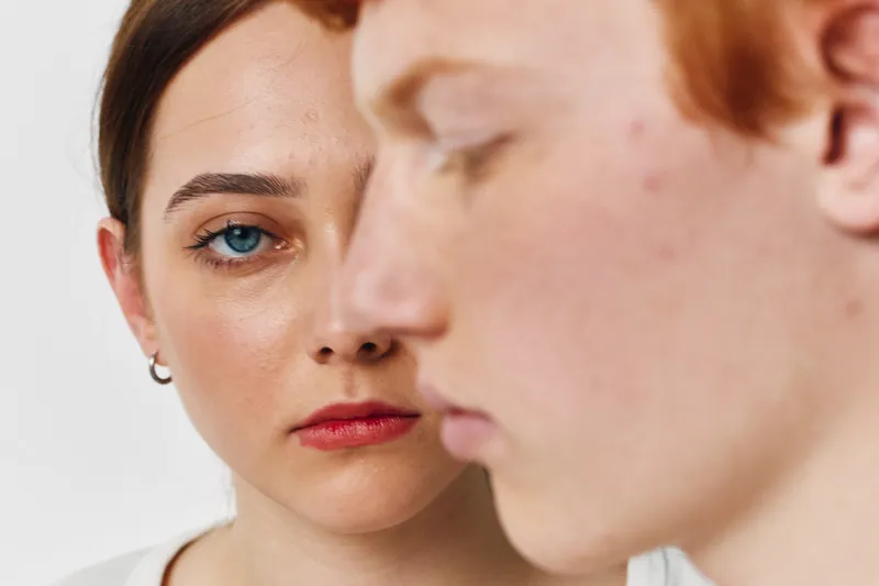 Portrait of young woman with blue eyes looking at redheaded man with serious expression, closeup of faces and skin details on white background. Intense gaze and emotion in studio setting.