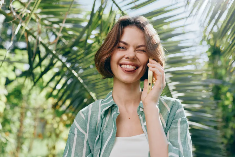 A joyful woman outdoors, smiling while talking on a mobile phone. Bright natural light and lush greenery create a fresh, relaxed lifestyle scene full of warmth and everyday connection.