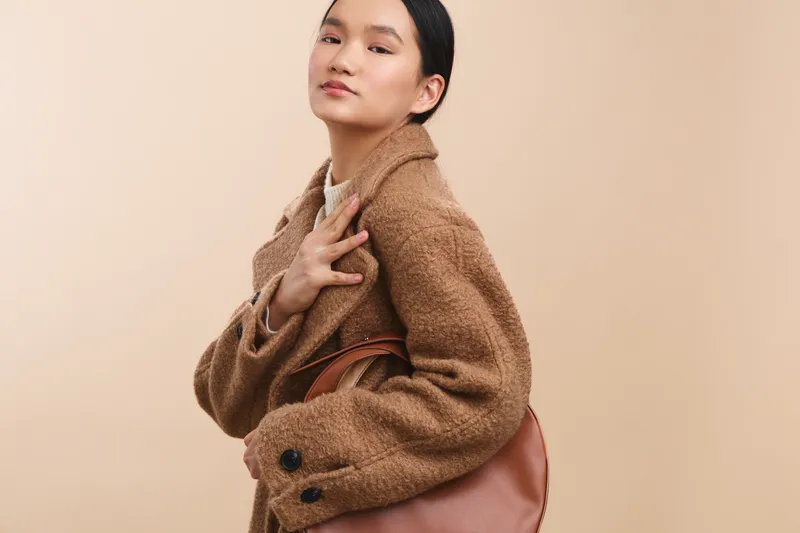Asian woman models a warm textured coat and soft handbag in a minimalist studio, exuding confidence and stylish elegance for a contemporary fashion shoot and attitude.