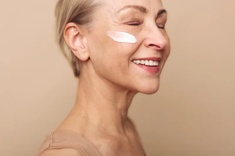 Smiling mature woman with short hair applying face cream on cheek, eyes closed in relaxed mood. Concept of skincare, aging beauty, self-care, and natural wellness in soft lighting.