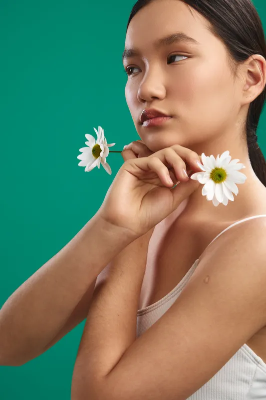 Asian woman holds daisies near her face against a vivid green backdrop, conveying calm elegance and natural beauty in a fresh, serene portrait with soft lighting.