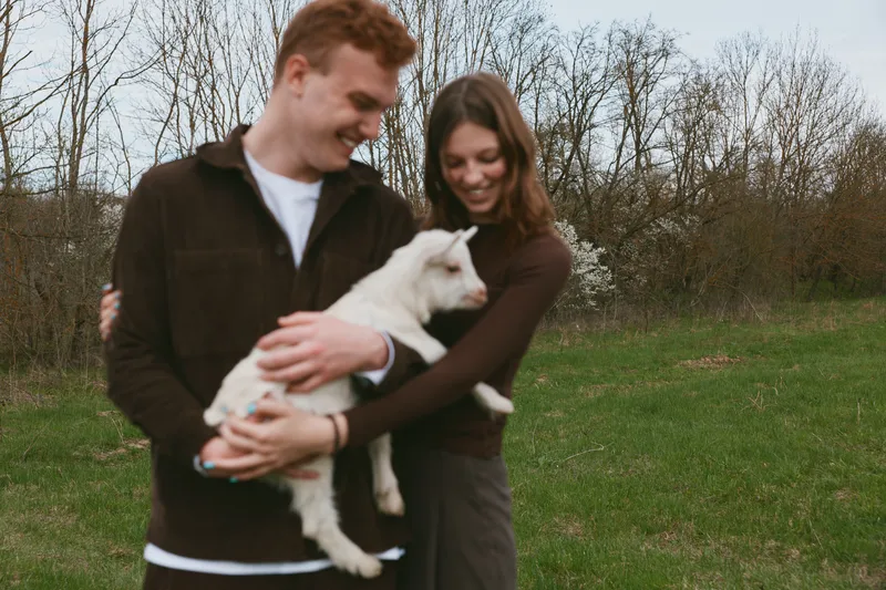 Joyful couple outdoors, holding a goat kid in their arms, smiling as the young animal nuzzles them. A lifestyle moment of connection, care, and rural happiness captured in natural light.