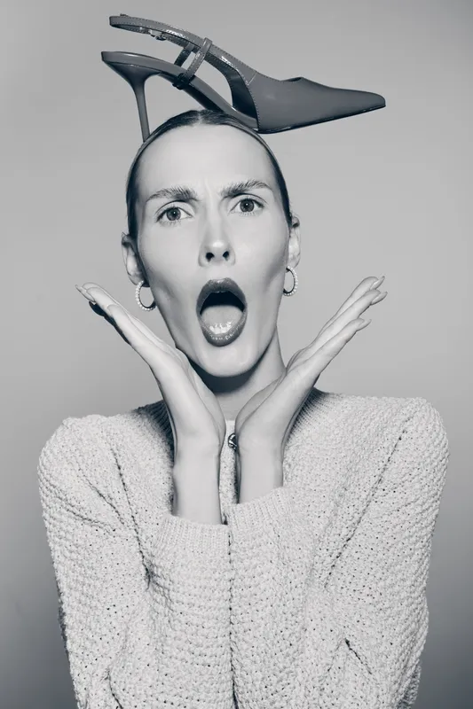 Surprised white woman with pale skin holds high heel shoe on head, wearing knitted sweater and hoop earrings on grey background.