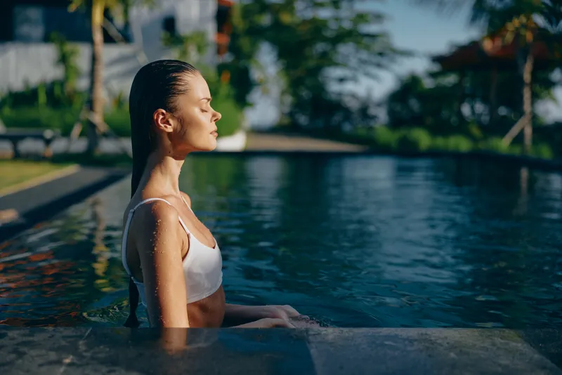 Young woman relaxes in a poolside on a sunny day, wearing a white bikini, posing calmly at the water edge, enjoying fresh air and solitude with a tropical garden backdrop