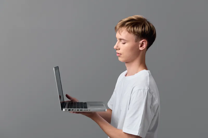 Boy in a white tee holds a laptop, focused on the screen, demonstrating digital learning and technology in a clean studio setup with a neutral background