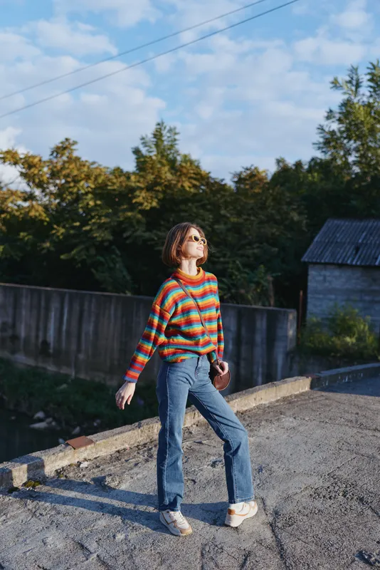 Young woman in a striped sweater and blue jeans stands outdoors by a wooden fence, wearing sunglasses and white sneakers, looking up and posing in golden hour casual outfit travel lifestyle scene.